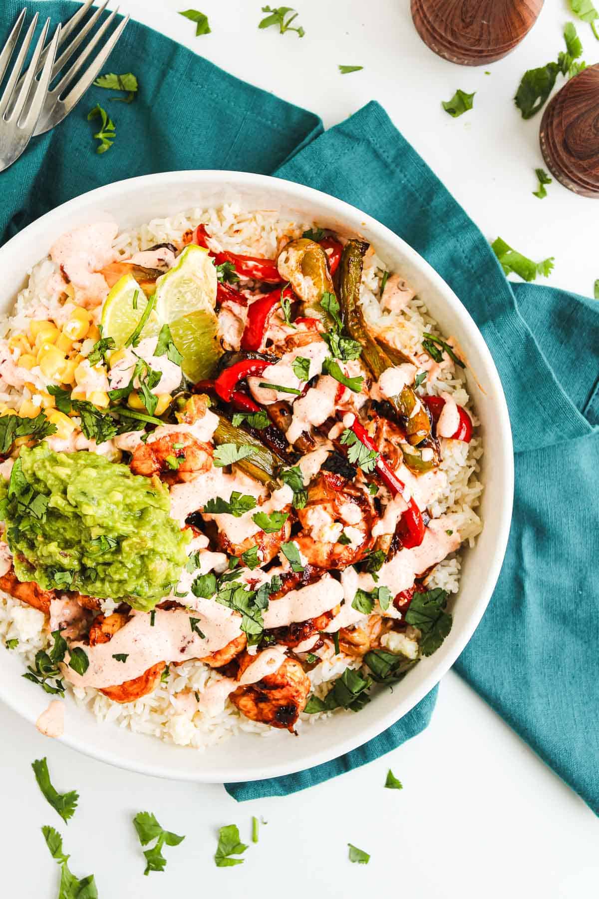 Bowl of rice topped with grilled chicken, veggies, guacamole, lime wedges, and creamy chipotle sauce, on a blue napkin&mdash;a delicious twist on classic fajita bowls.