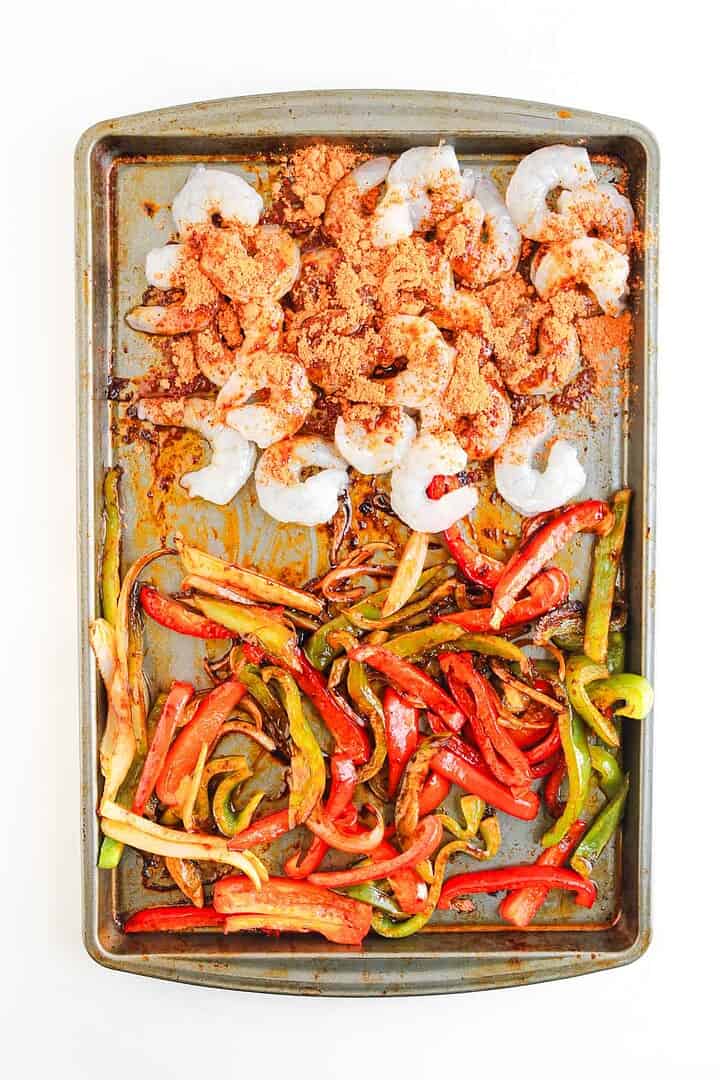 Sheet pan with raw shrimp topped with seasoning and mixed sliced red, yellow, and green bell peppers&mdash;perfect for building flavorful Shrimp Fajita Bowls served with a zesty chipotle sauce.