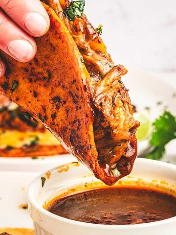 A hand dips a birria taco filled with meat into a cup of broth, with cilantro and lime in the background.