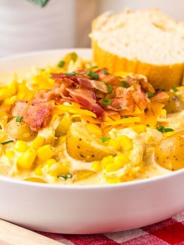 A bowl of creamy potato corn chowder topped with bacon and cheese, served with bread on a red plaid cloth.