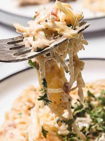 A fork lifts creamy pasta above a plate garnished with fresh herbs.