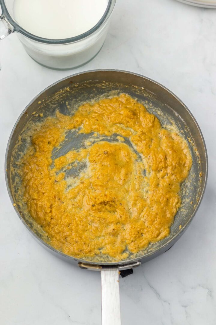 A pan with a thick, golden-yellow roux mixture sits on a white countertop near a measuring cup of milk.
