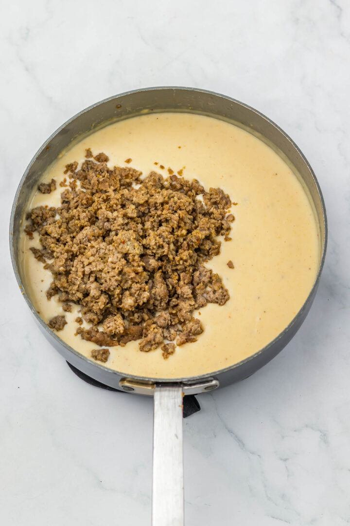 A skillet with creamy cheese sauce and cooked ground sausage—perfect for making Sausage Biscuit Gravy casserole—sits on a white marble surface.