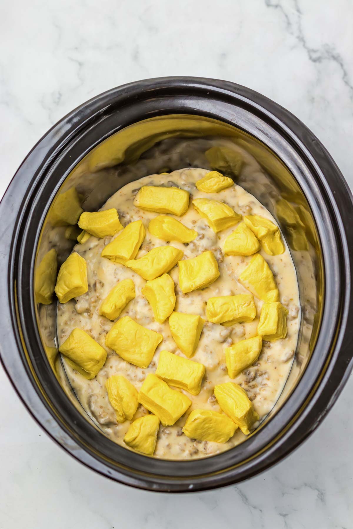 Crockpot filled with creamy sausage biscuit gravy mixture, topped with chunks of yellow biscuit dough on a marble surface.