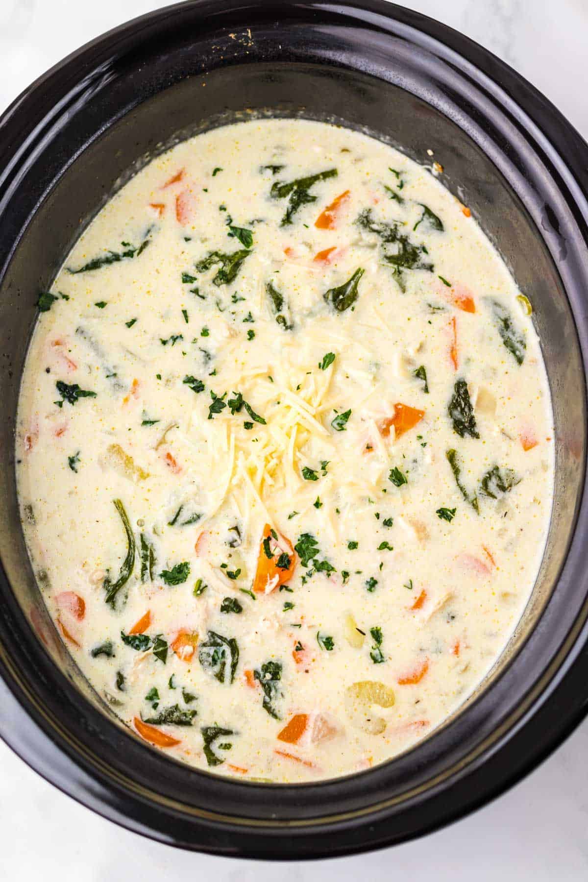 Creamy chicken gnocchi soup with vegetables and spinach, topped with shredded cheese, simmering in a black slow cooker.