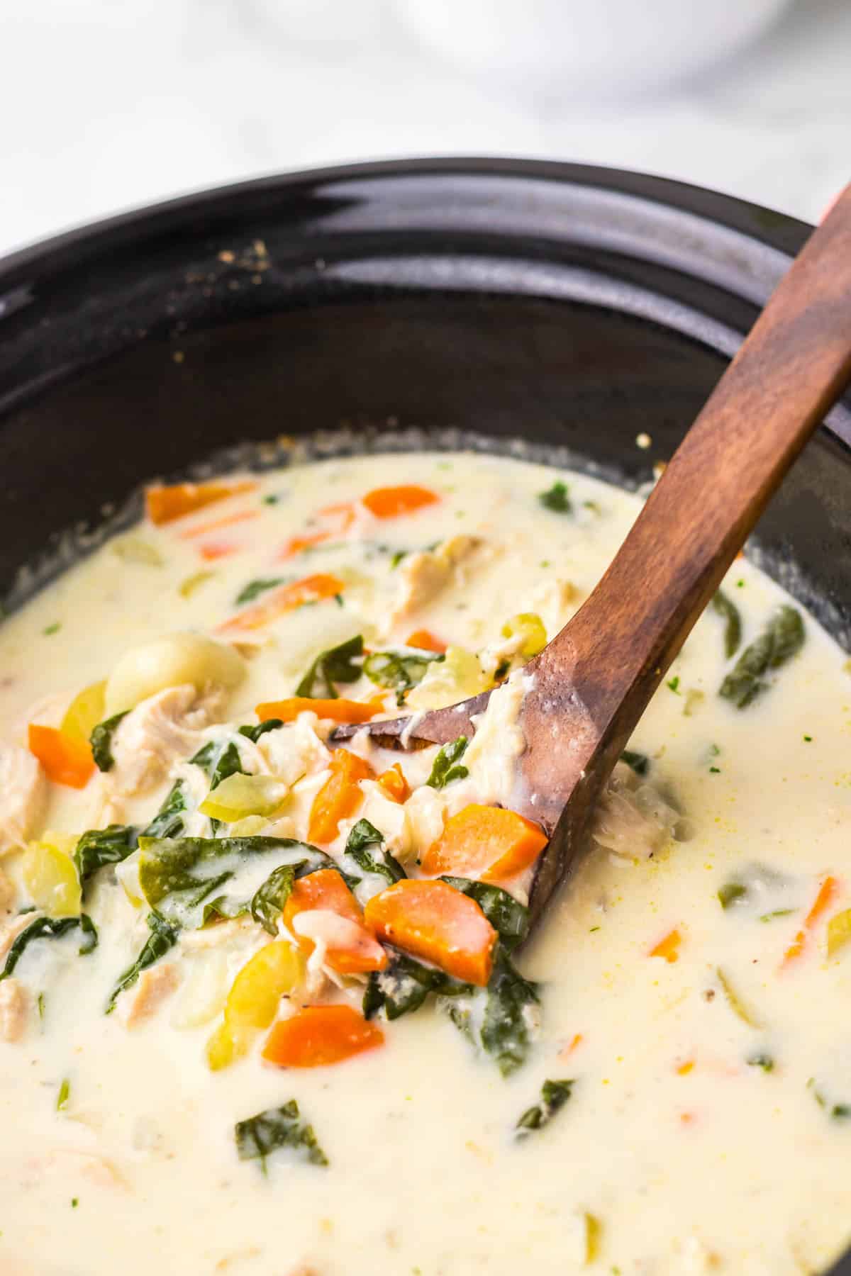 A wooden spoon stirs creamy chicken and vegetable soup in a black slow cooker.