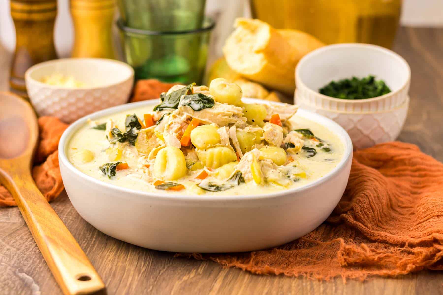 A bowl of creamy Chicken Gnocchi Soup with vegetables sits on a wooden table, accompanied by fresh bread and bowls&mdash;perfect comfort food, especially when made in a slow cooker.