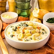 Bowl of creamy chicken gnocchi soup with tender vegetables, served alongside bread and green glassware.