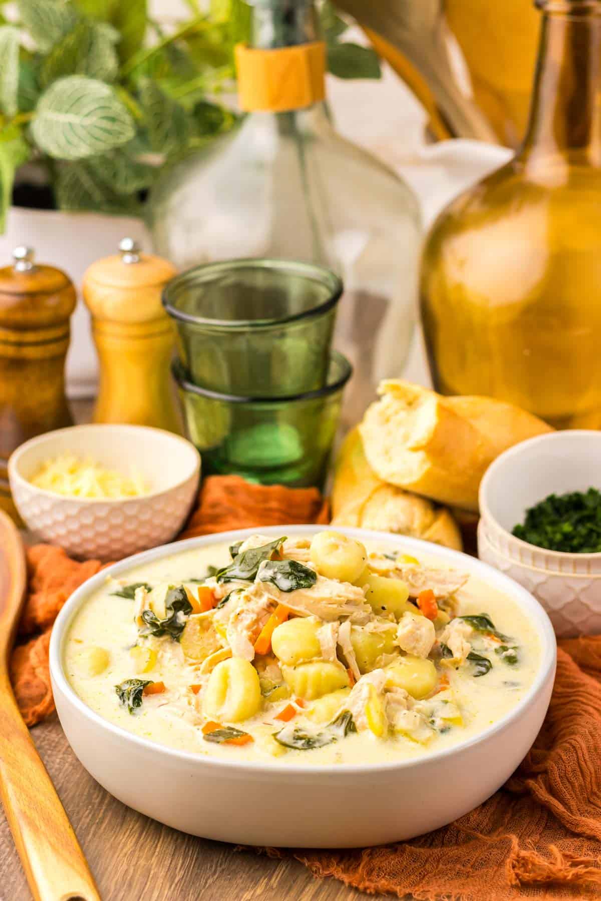 Bowl of creamy chicken gnocchi soup with tender vegetables, served alongside bread and green glassware.