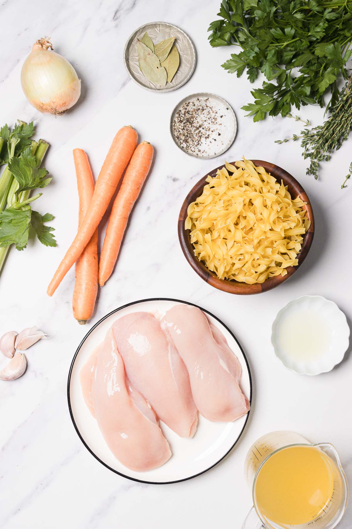 Raw chicken breasts, uncooked egg noodles, carrots, celery, onion, garlic, herbs, and seasonings on a white surface.