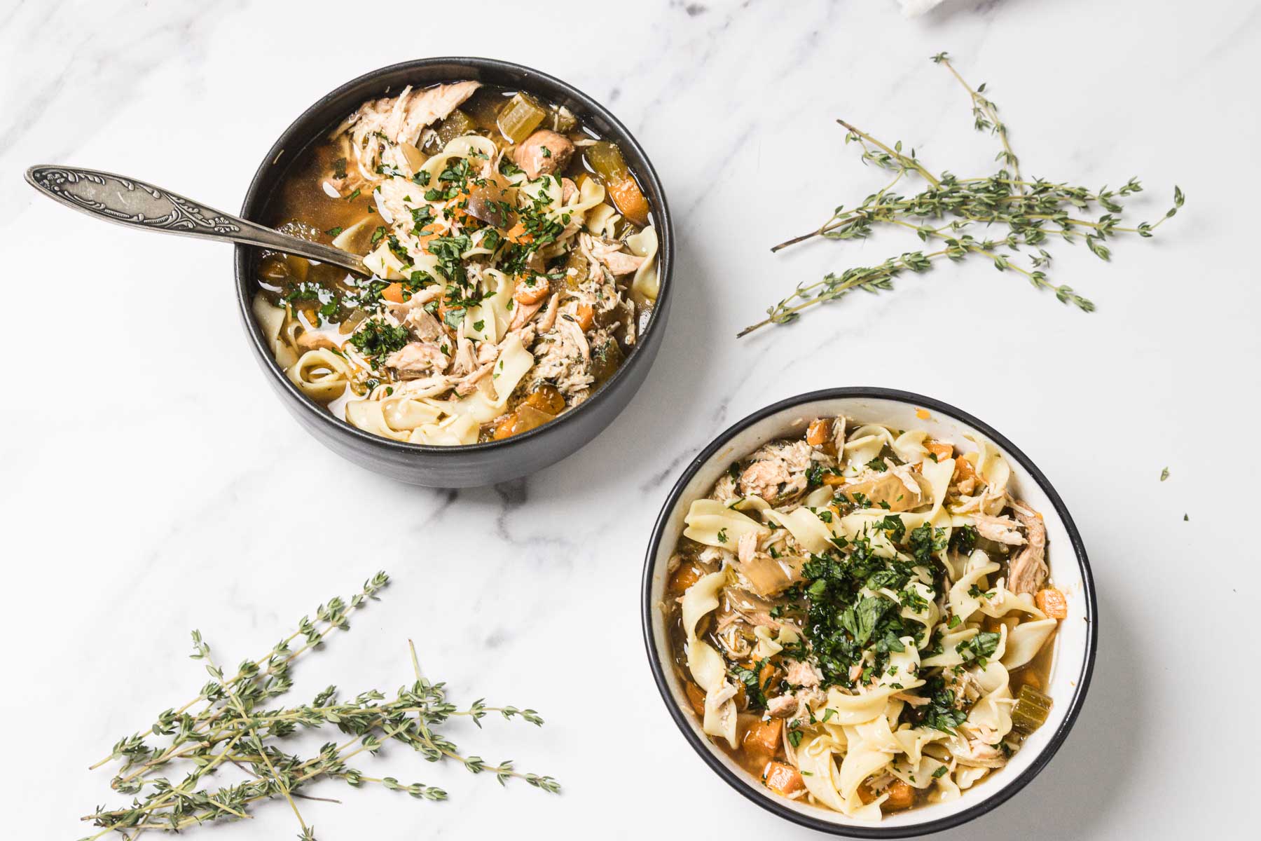 Two bowls of comforting Slow Cooker Chicken Noodle Soup with herbs sit on a white surface, garnished with fresh thyme sprigs nearby.