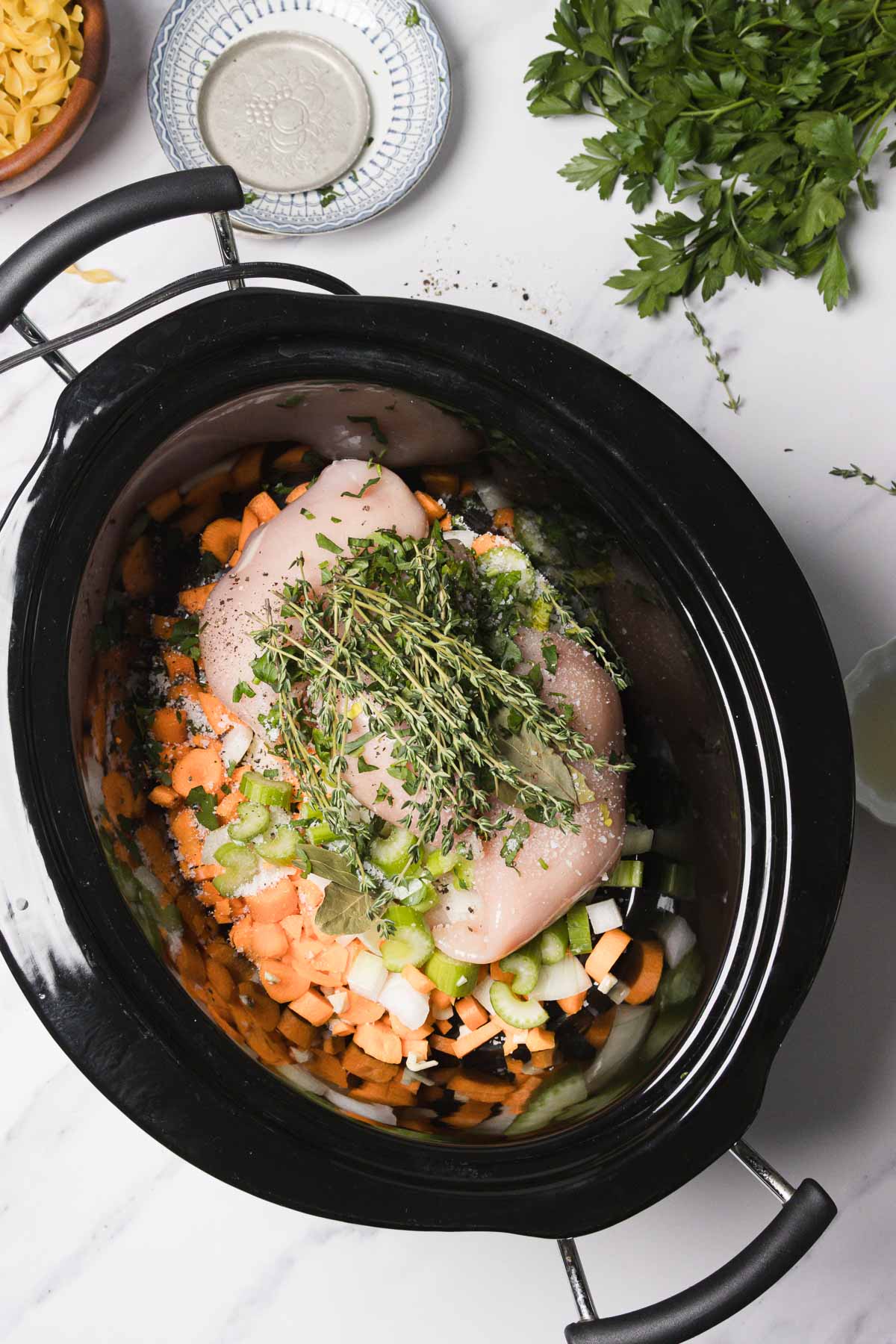 Raw chicken breasts, chopped carrots, celery, onions, and fresh herbs in a slow cooker on a white countertop&mdash;perfect ingredients to start a comforting Chicken Noodle Soup recipe.