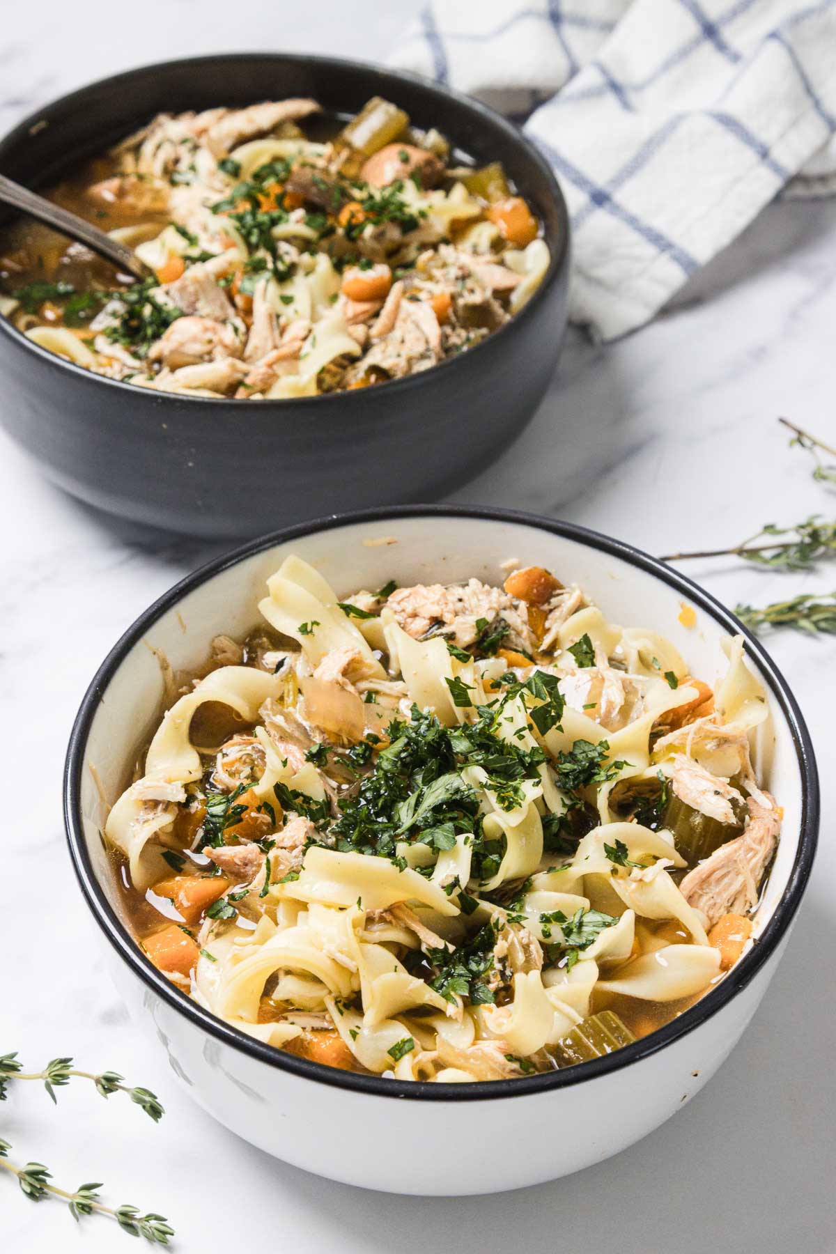 Two bowls of Chicken Noodle Soup with wide noodles, vegetables, and chopped herbs on top sit on a white surface&mdash;perfect for trying a cozy slow cooker recipe at home.