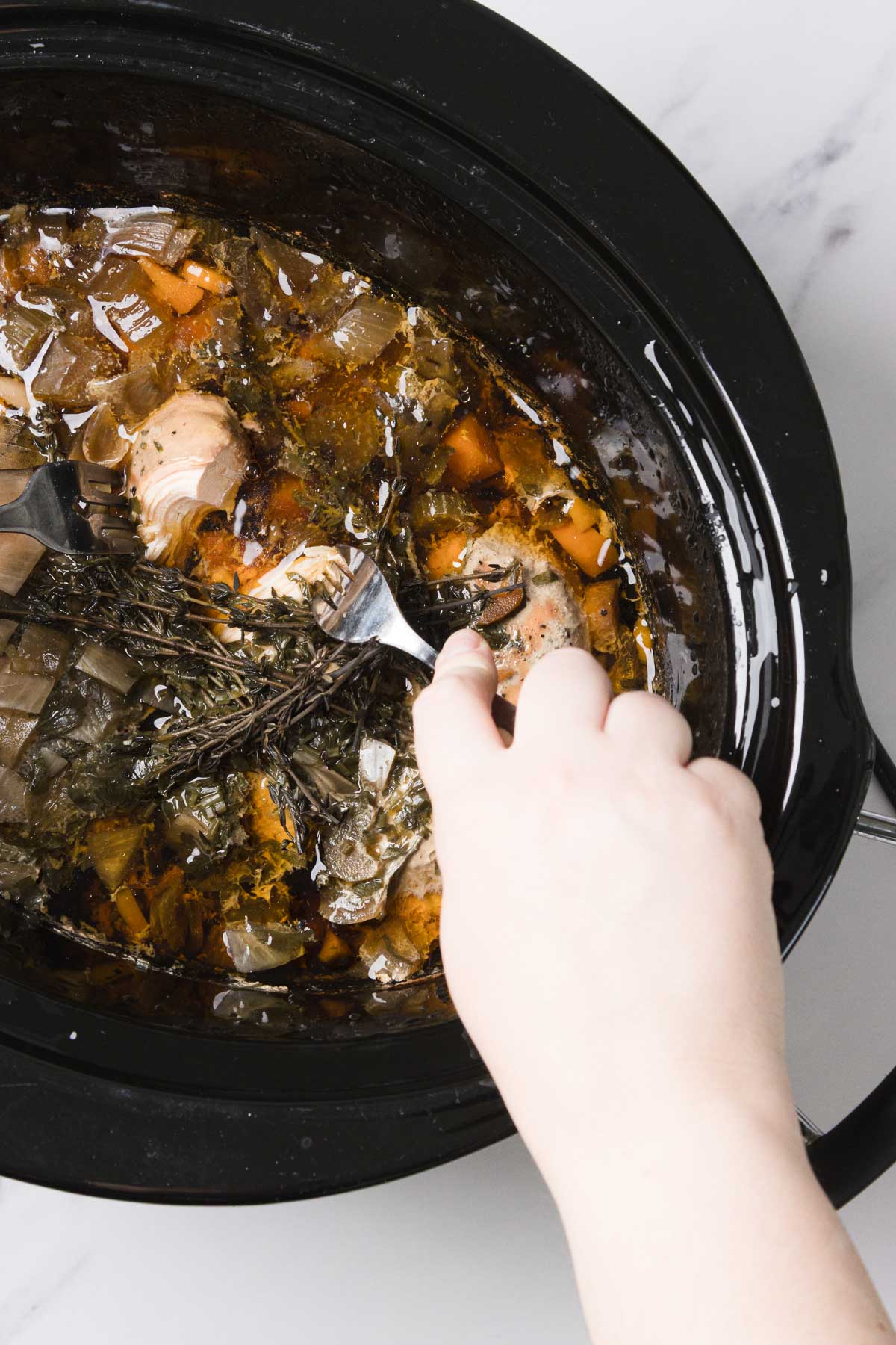 A hand uses a fork to check cooked food in a slow cooker with herbs, vegetables, and broth for a comforting Chicken Noodle Soup recipe.