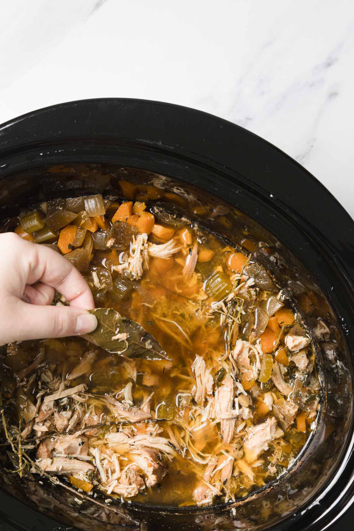 Hand removing a bay leaf from shredded meat and vegetables in a slow cooker, perfect for a comforting Chicken Noodle Soup recipe with rich broth.