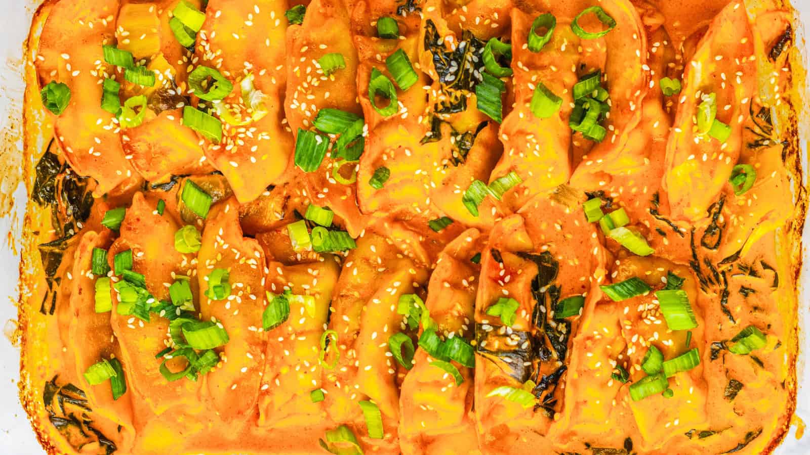 Close-up of baked dumplings in orange sauce, topped with chopped green onions and sesame seeds.