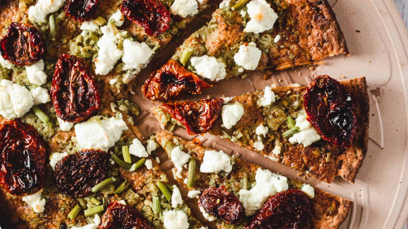 A close-up of a pizza topped with sun-dried tomatoes, goat cheese, and green vegetables, cut into slices.