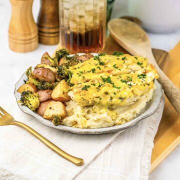 A plate of chicken with sauce, mashed potatoes, and roasted vegetables sits on a table with a drink.