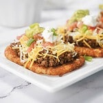 Two tostadas topped with seasoned ground beef, shredded cheese, salsa, guacamole, sour cream, and green onions.