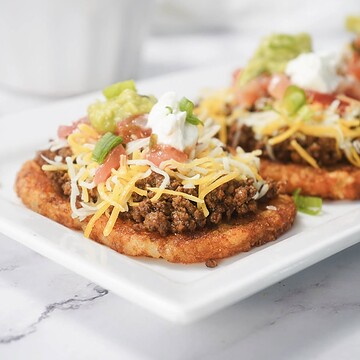 Two tostadas topped with seasoned ground beef, shredded cheese, salsa, guacamole, sour cream, and green onions.