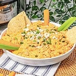 A bowl of creamy Buffalo Chicken Dip topped with green onions, served with chips, celery, and carrot sticks—perfect for any Crockpot party spread.