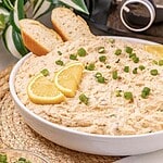 A bowl of creamy crab dip topped with lemon slices and chopped green onions, served with bread slices—a delicious recipe perfect for your next gathering.