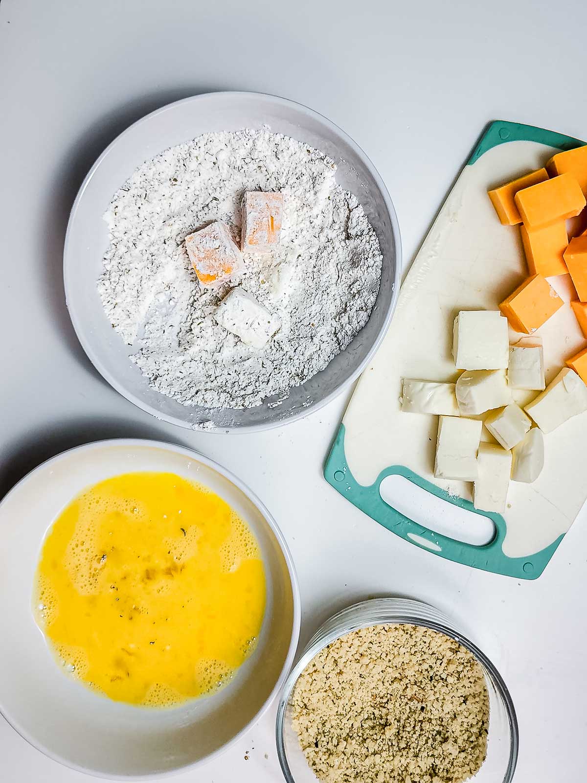 Bowls with flour-coated cheese bites, beaten eggs, and breadcrumbs ready for the air fryer sit next to a cutting board piled with cubed cheese&mdash;perfect for making crispy snacks.