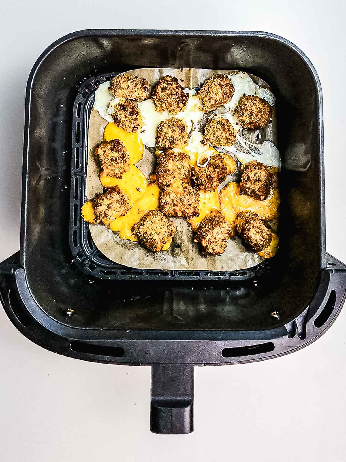 Cooked meatballs and gooey cheese bites create a crispy finish in an air fryer basket lined with parchment paper.