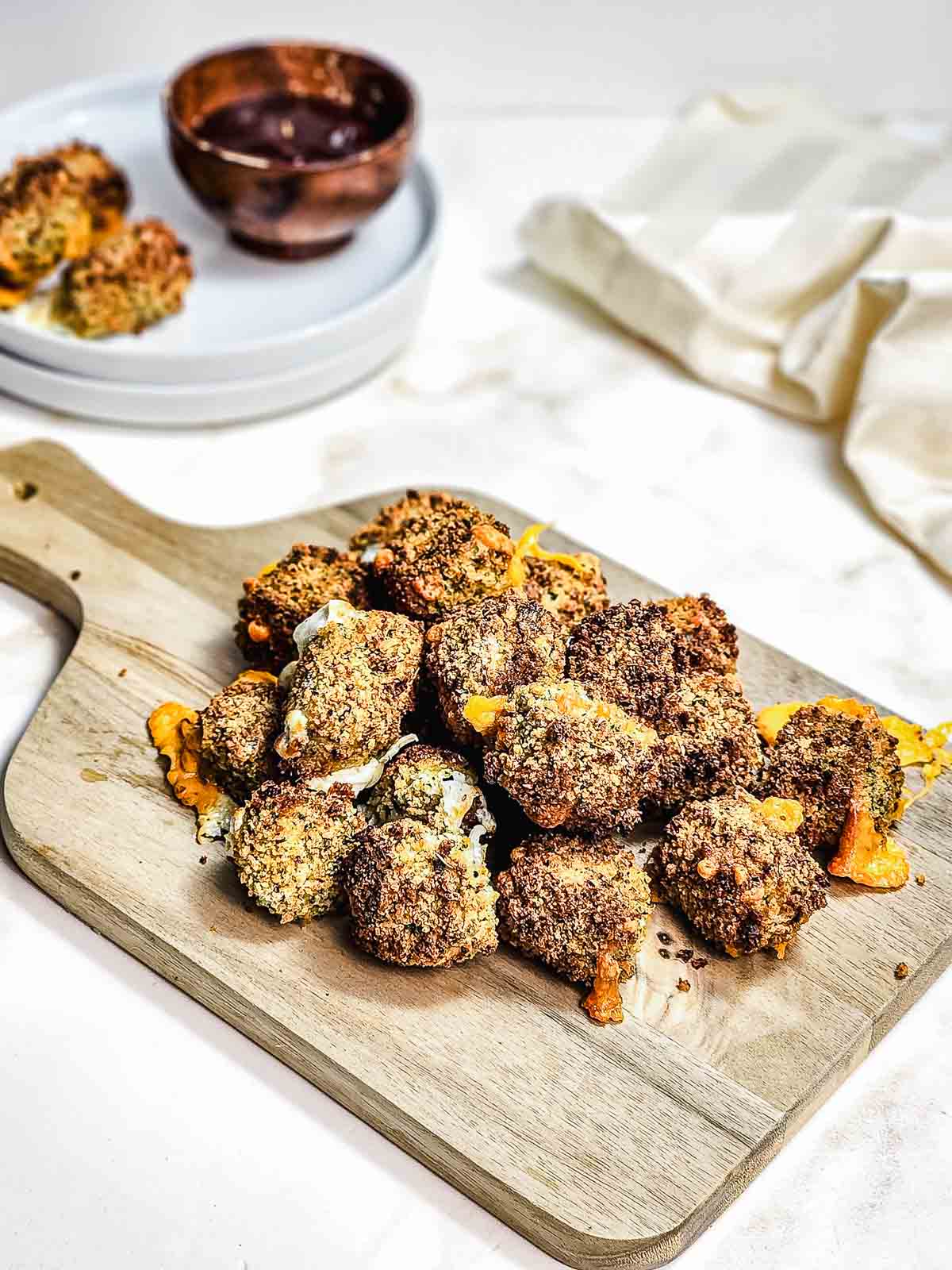 A wooden board with crispy, breaded broccoli bites, baked to perfection in the air fryer, with a plate and dipping sauce in the background.