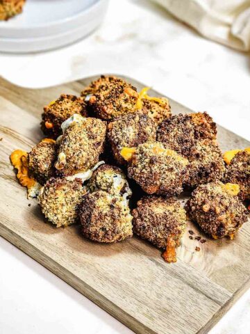 Crispy, breaded broccoli and cheese bites served on a wooden board, with sauce in a bowl and a plate in the background—perfect for making in your air fryer.