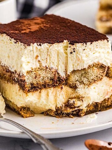 A slice of creamy tiramisu on a plate with a fork, with coffee cups and more tiramisu in the background.