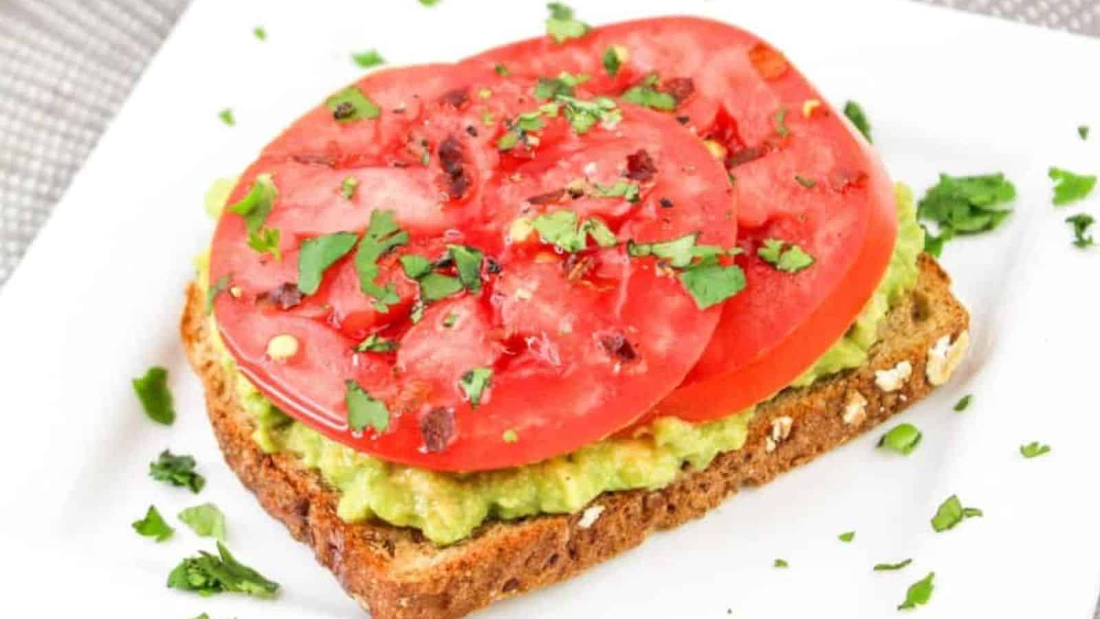 A slice of whole-grain bread with avocado spread topped with two tomato slices and garnished with chopped cilantro on a white square plate.