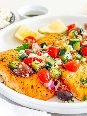 Grilled salmon topped with a Greek salad of tomatoes, cucumbers, olives, and feta, served with lemon wedges.