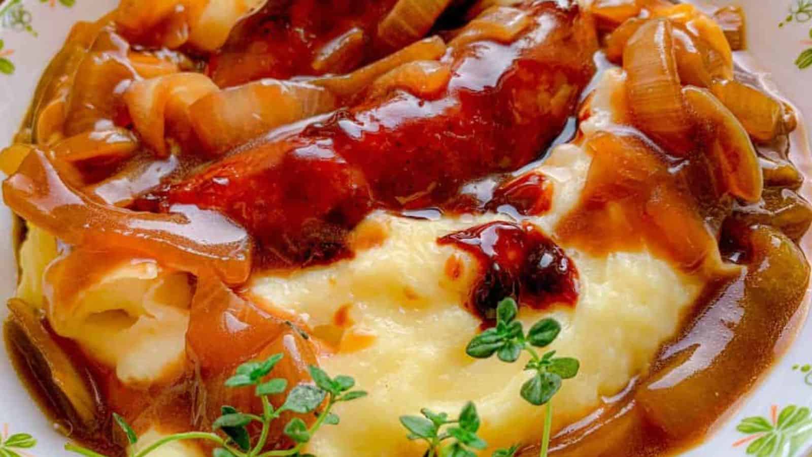 A plate of mashed potatoes topped with sausages and caramelized onions in a rich brown gravy. A sprig of fresh thyme garnishes the dish. The plate sits on a green patterned cloth.