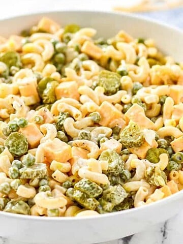 Bowl of creamy macaroni salad with peas, diced cheese, and pickles on a marble surface with a wooden spoon nearby.