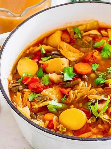 A pot of vegetable soup with carrots, cabbage, tomatoes, and herbs surrounded by ingredients on a white table.