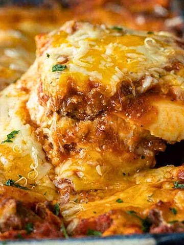 A slice of cheesy, baked lasagna being lifted from a pan, topped with herbs and melted cheese.