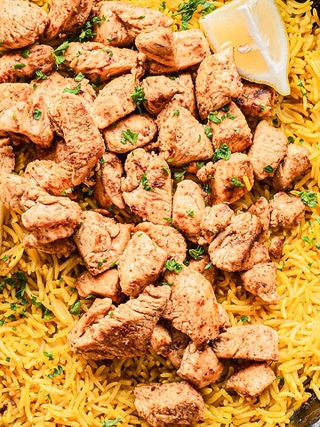 A skillet with yellow rice, spiced chicken pieces, and two lemon wedges on top.