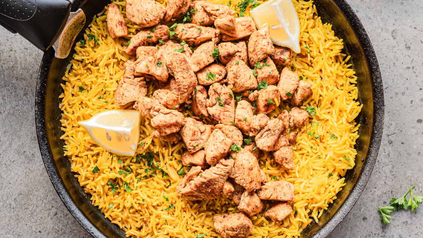 A fork in a bowl of yellow rice with grilled chicken, lemon wedges, herbs, and a dollop of white sauce.