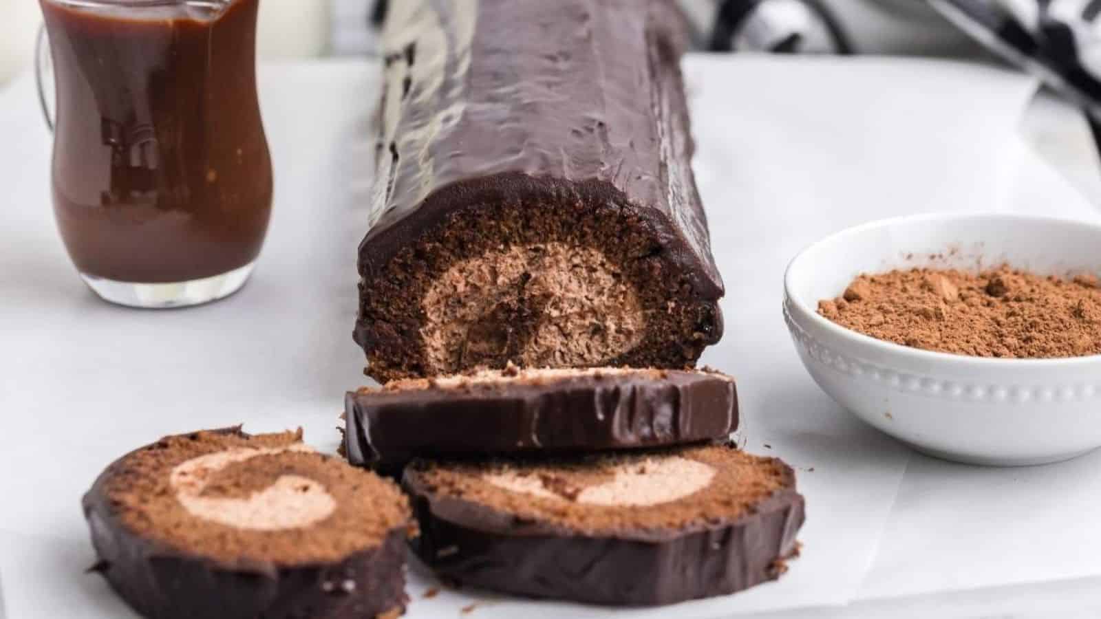Chocolate swiss roll with three slices cut, revealing cream filling. A small bowl of cocoa and a jug of chocolate sauce are beside it on a white surface.