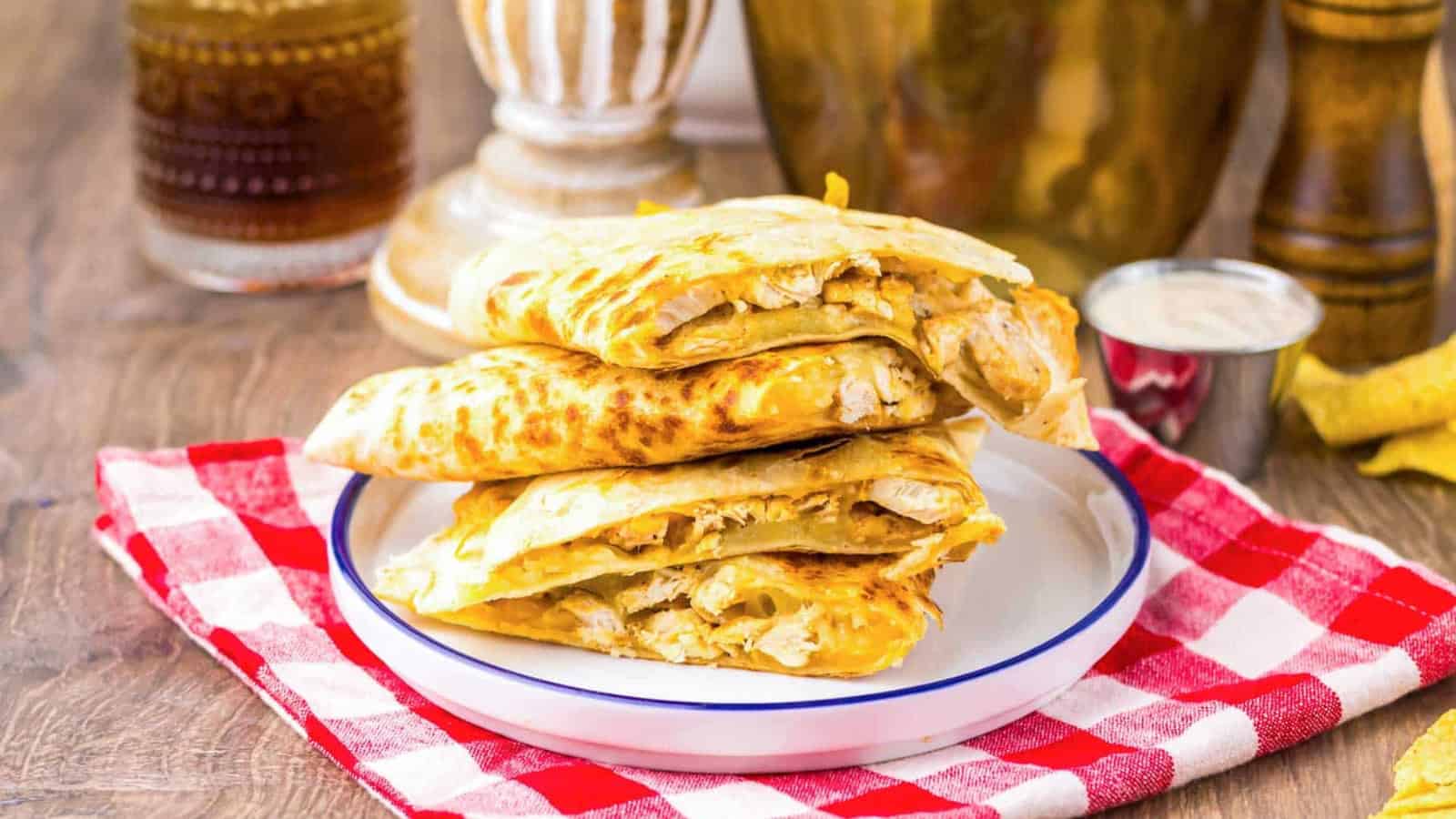 Three pieces of stuffed flatbread stacked on a plate, on a red checkered cloth, with sauce on the side.