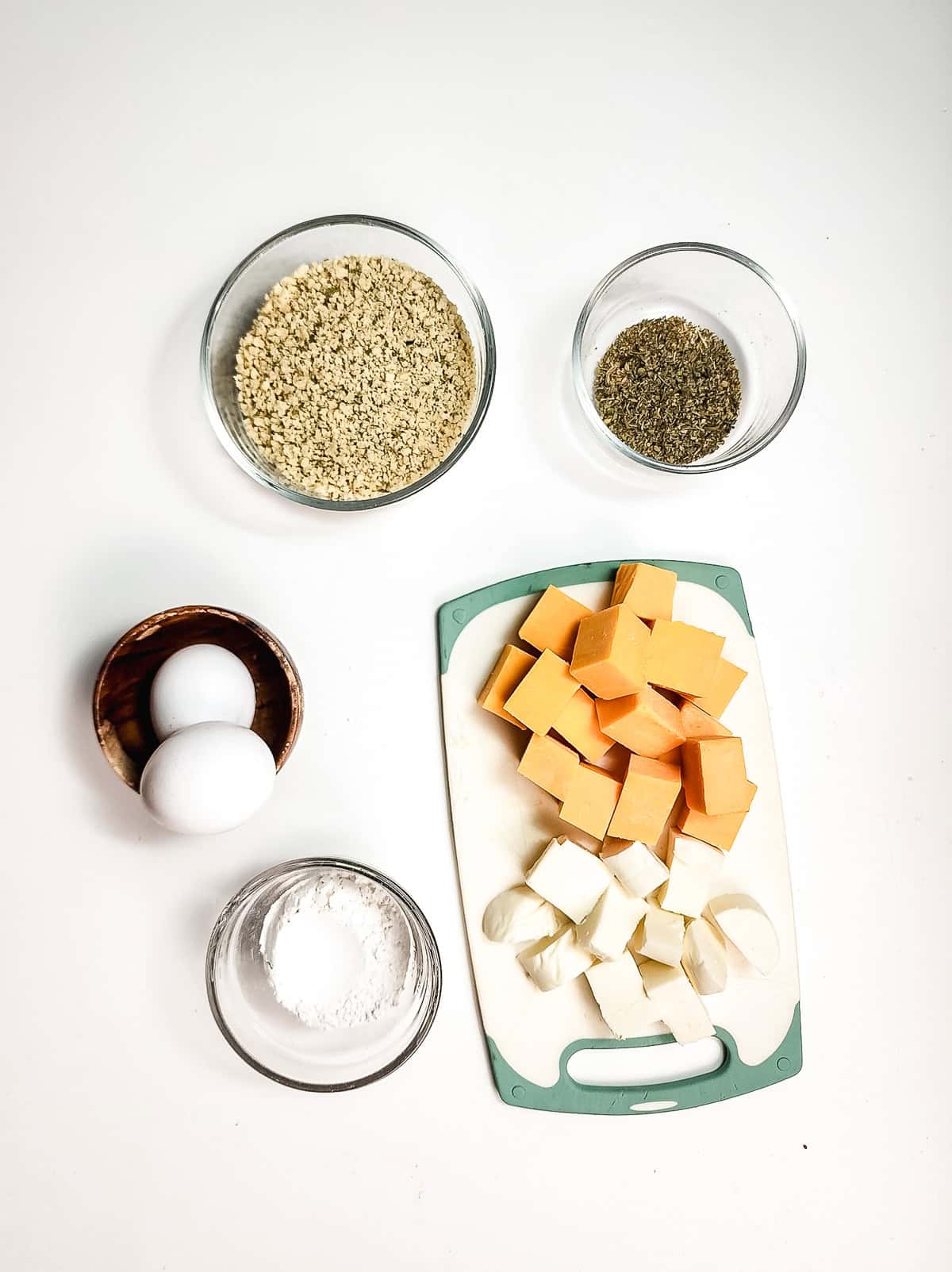 Cubes of cheese and squash, two eggs, flour, oats, and herbs in bowls on a white background&mdash;everything you need to make crispy air fryer cheese bites.