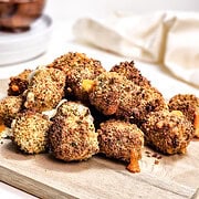 A pile of breaded and baked cauliflower bites on a wooden board, reminiscent of crispy air fryer cheese bites, with a white cloth and bowls in the background.