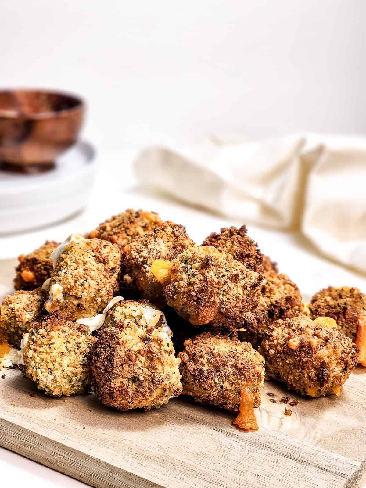 A pile of breaded and baked cauliflower bites on a wooden board, reminiscent of crispy air fryer cheese bites, with a white cloth and bowls in the background.