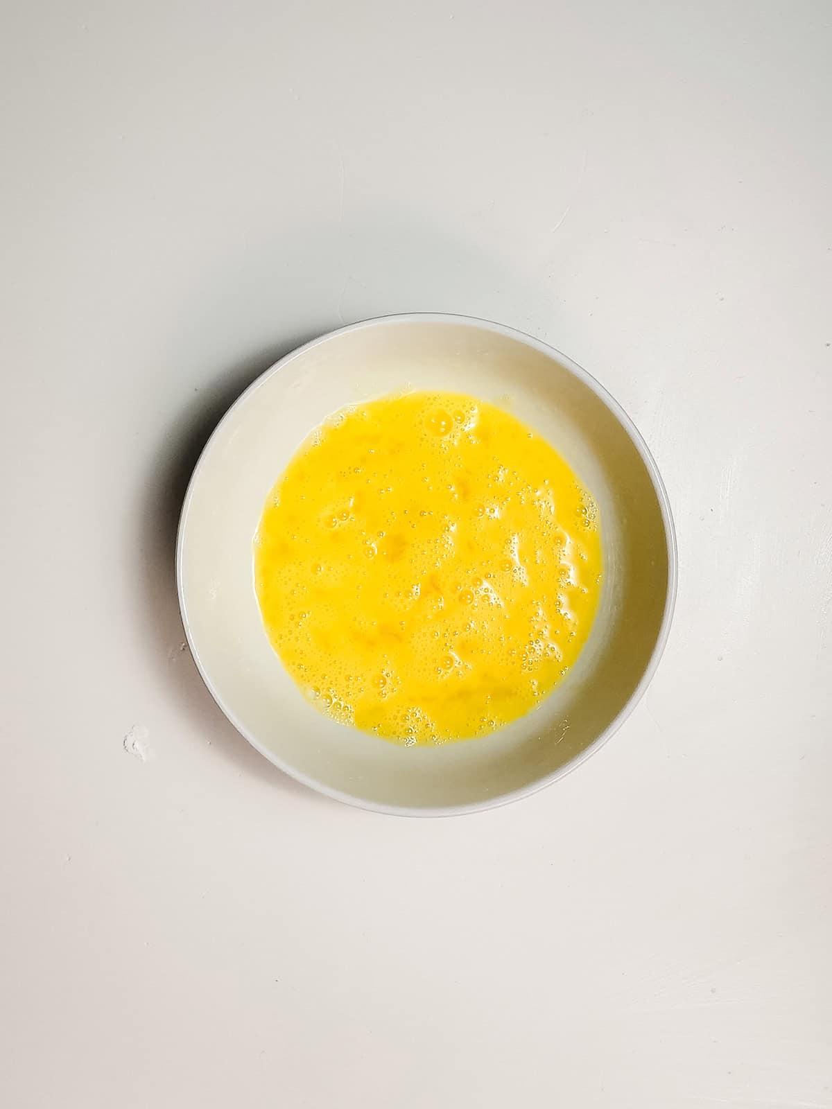 A white bowl filled with beaten eggs sits on a plain white surface, ready to coat crispy air fryer cheese bites before cooking.