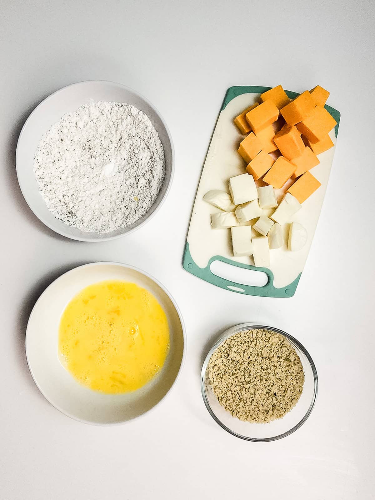 Four bowls with flour, whisked eggs, breadcrumbs, and a cutting board with cubed sweet potato and onion&mdash;perfect for prepping crispy air fryer cheese bites.