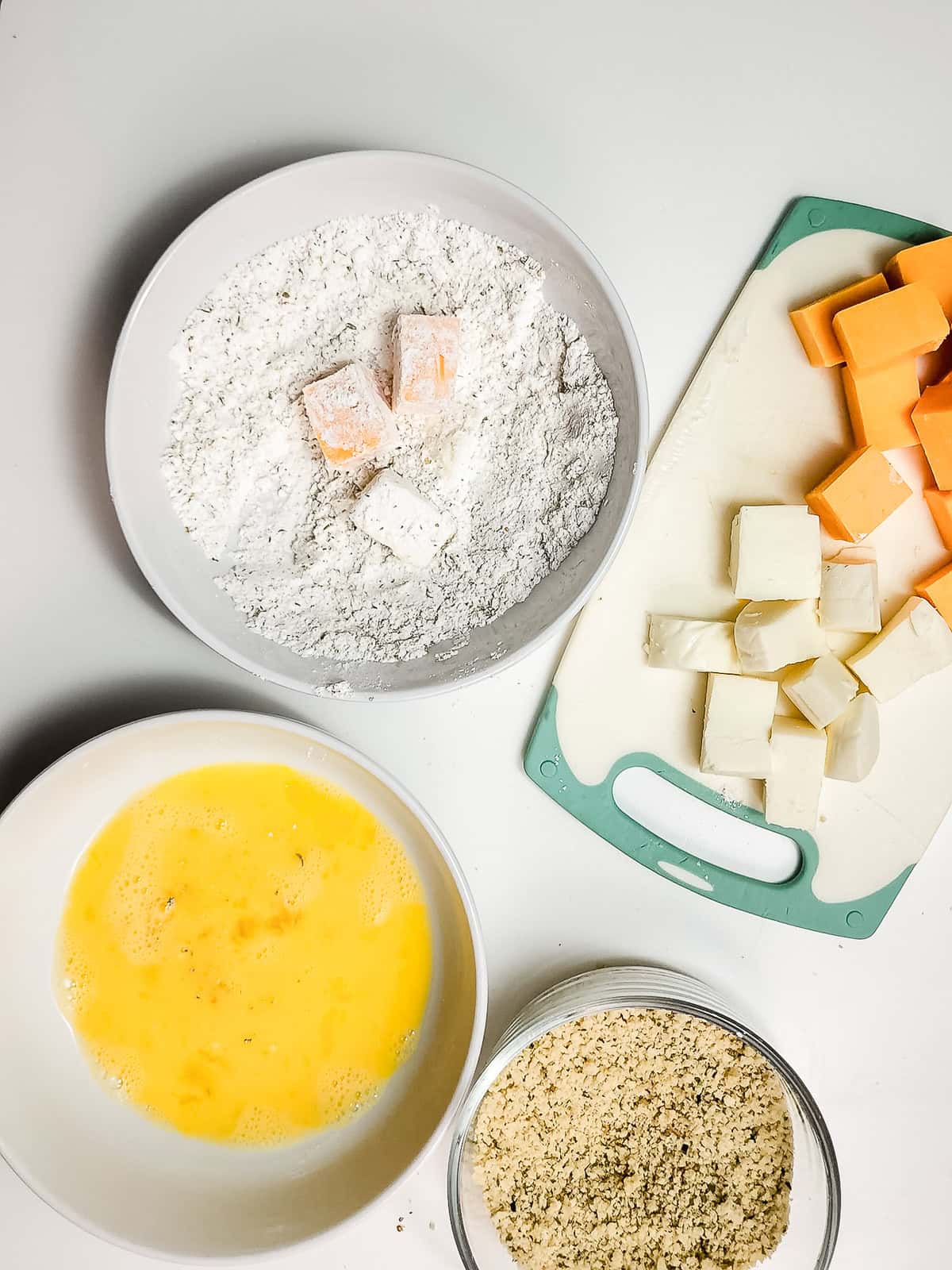 Bowls of flour with butter, beaten eggs, breadcrumbs, and a cutting board with cubed cheese on a white surface&mdash;everything you need to make crispy air fryer cheese bites.