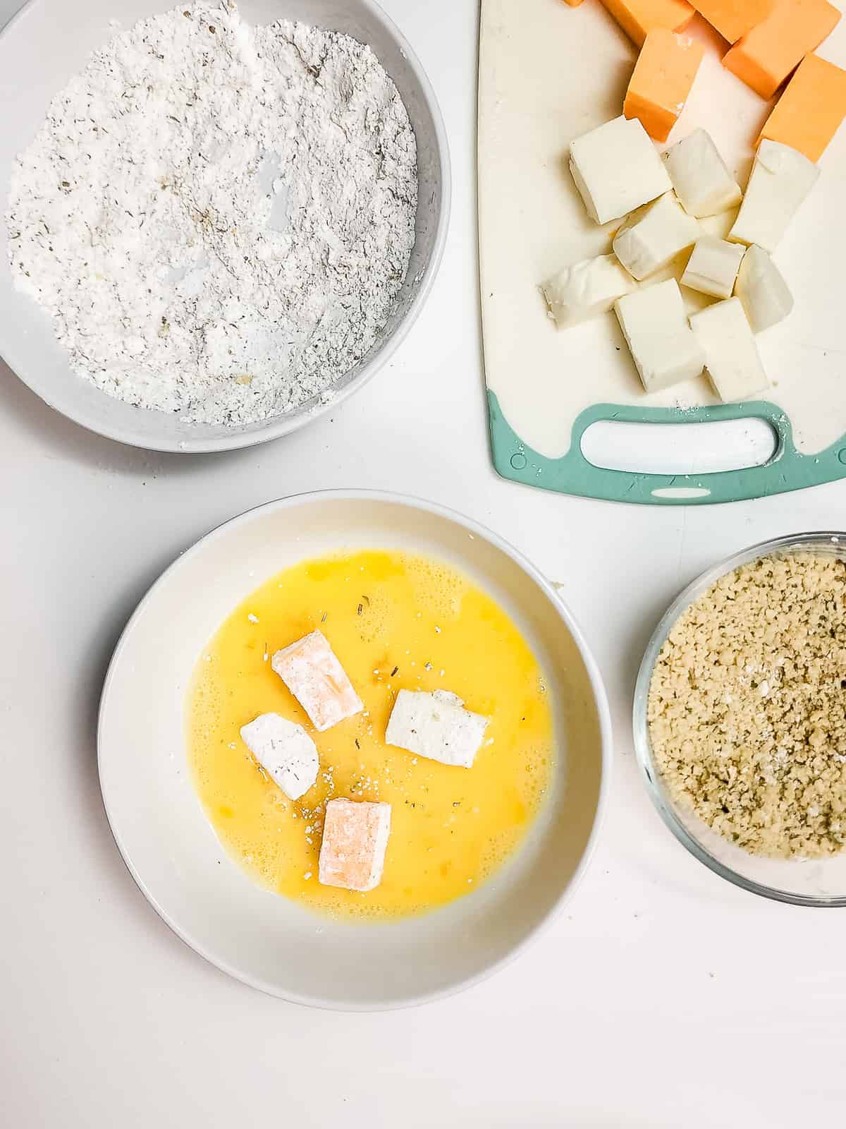 Bowls of flour, egg wash with cheese cubes, and breadcrumbs sit next to a cutting board with cubed cheese, ready for making crispy air fryer cheese bites.