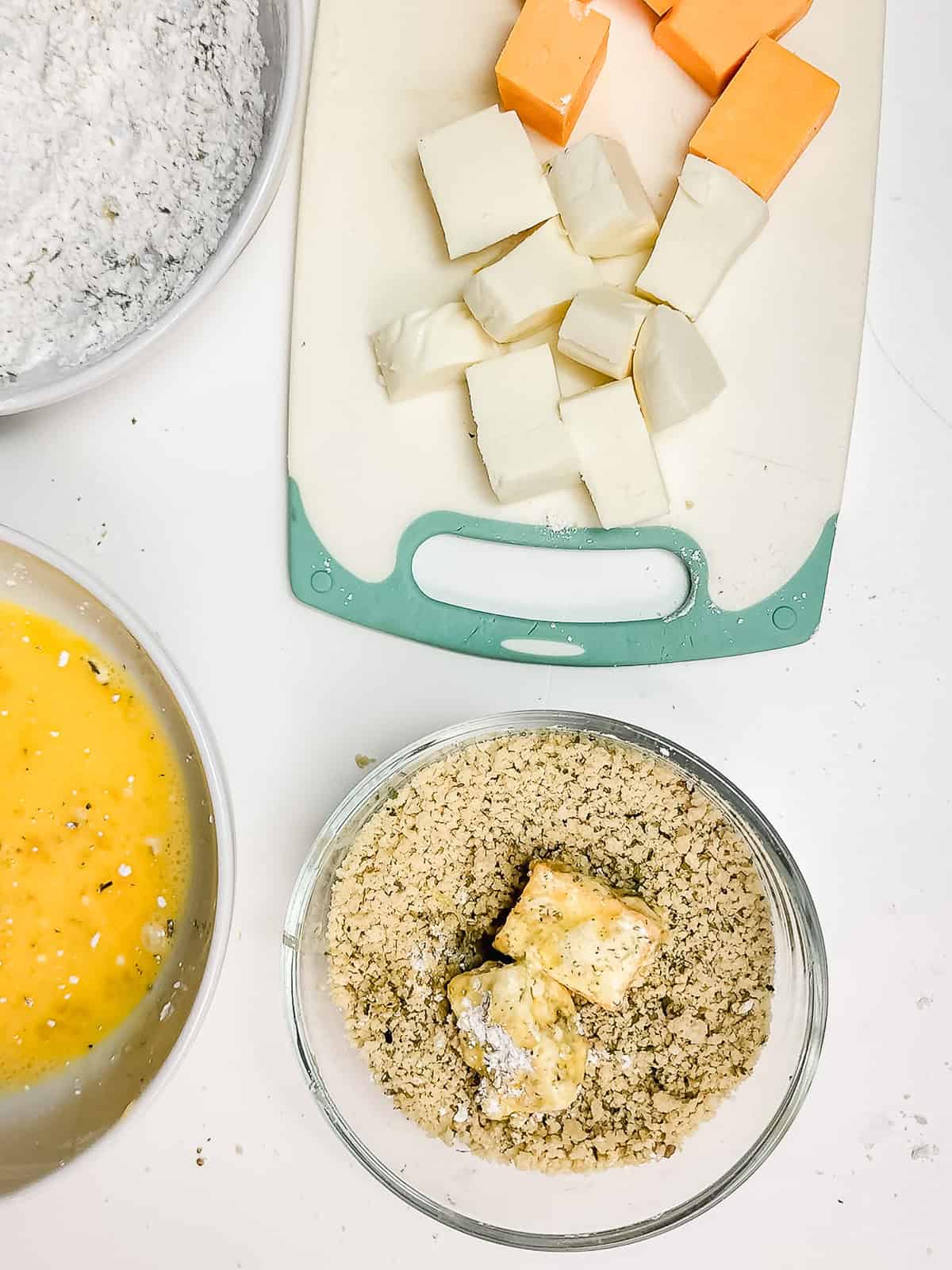 Cubed cheese on a cutting board, with bowls of flour, beaten eggs, and breadcrumb mixtures nearby&mdash;perfect for making crispy air fryer cheese bites.