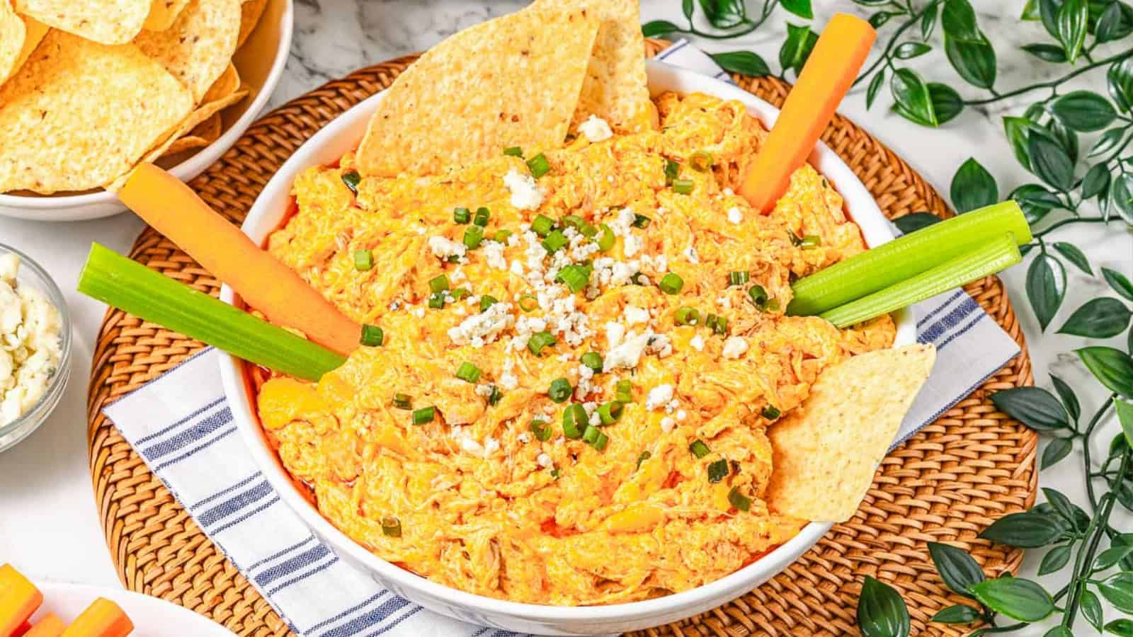 A bowl of buffalo chicken dip garnished with celery sticks, carrot sticks, chips, and sprinkled cheese.